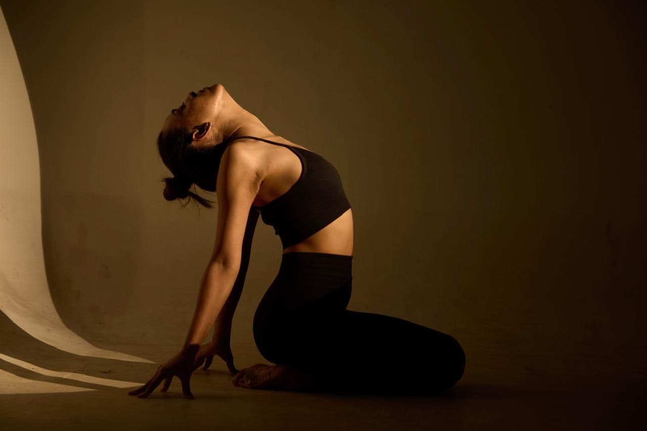 A woman kneeling in a deep yoga stretch, lit warmly against a studio wall