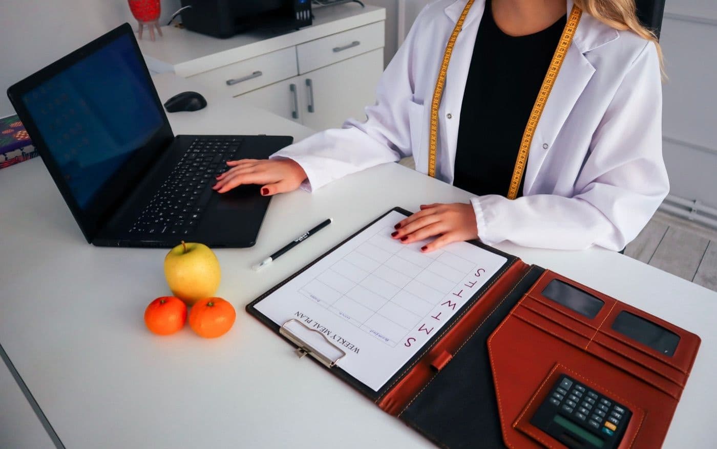 A nutritionist at a desk with a laptop, a weekly meal-plan sheet and fresh fruit